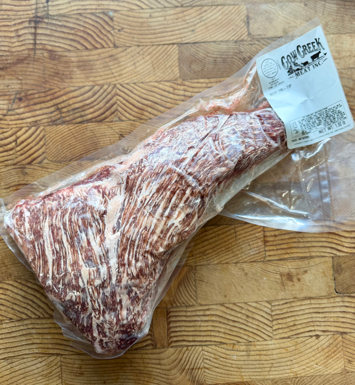 A vacuum-sealed raw cut of marbled tri tip meat with a white label reading Cow Creek Meat Inc., placed on a wooden cutting board. Size of meat is 1.92 lbs