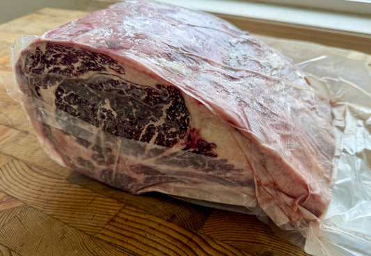 A large frozenmarbled cut of meat, likely a ribeye roast, sealed in a clear plastic vacuum bag and resting on a wooden cutting board.